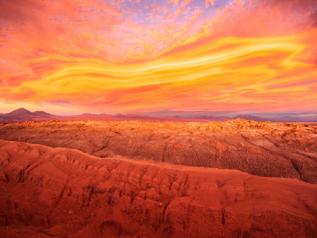 Paisaje desértico de horizonte amplio bajo un cielo despejado. Terreno árido con tonalidades ocres y marrones, presencia de formaciones rocosas o dunas de arena. Al fondo, siluetas de montañas o volcanes bajo una iluminación intensa de luz natural.