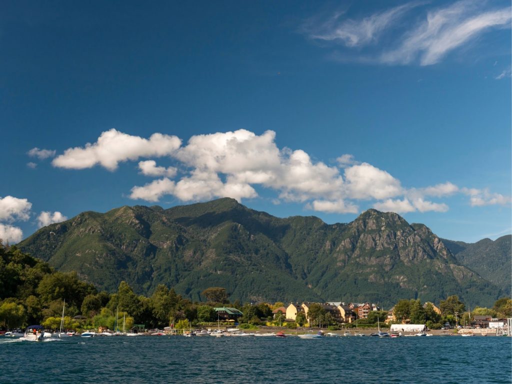 Paisaje del sur de Chile que combina un volcán de cono prominente y cima nevada con un gran cuerpo de agua (lago) en la base. El entorno incluye bosques densos de árboles nativos y una costa con arena de tonos oscuros.