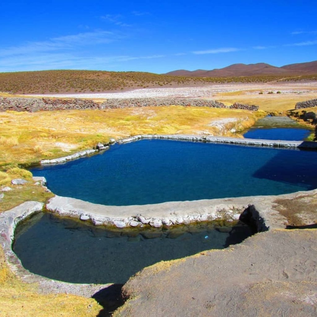 Vista superior de piscinas com água azul nas Termas de Enquelga. Situam-se numa turfeira alto-andina dourada com muros de pedra tradicionais, em frente a um vale desértico e vulcões no horizonte destas termas do norte do Chile.