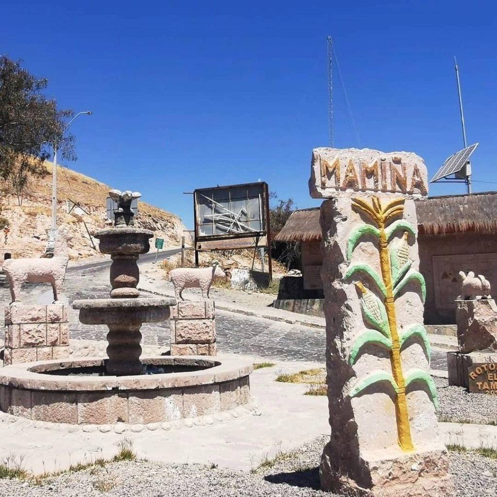 Primeiro plano do tótem de pedra à entrada de Mamiña, que mostra uma espiga de milho pintada em verde e amarelo e a palavra "MAMIÑA". Ao fundo, uma fonte de água tradicional de três níveis e a paisagem desértica.