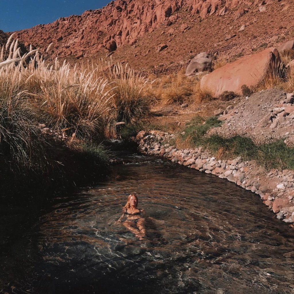 Uma mulher de costas flutua numa poça de água cristalina das termas de Puritama, rodeada de arbustos altos e grandes blocos de rocha avermelhada num desfiladeiro soalheiro perto de San Pedro de Atacama.