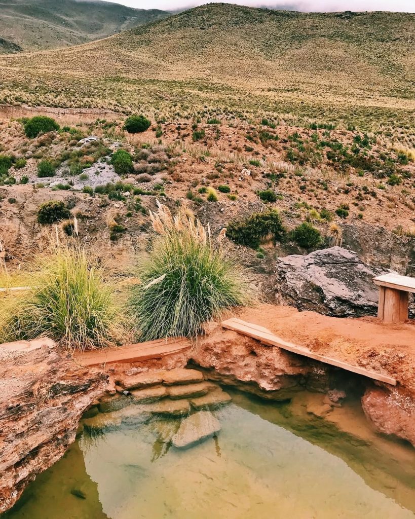 Vista em ângulo da poça rústica das Termas de Jurasi, umas das termas do norte do Chile, com degraus de pedra. Ao fundo, uma encosta andina coberta de vegetação baixa e o céu nublado.
