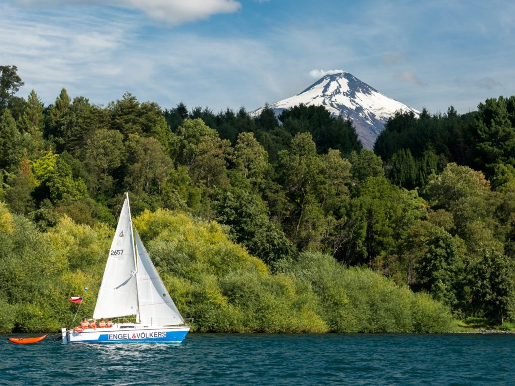 Un velero blanco navega sobre las aguas azules del Lago Villarrica frente a una densa cortina de bosque nativo, coronado al fondo por la imponente cumbre nevada del volcán Villarrica bajo un cielo semidespejado.