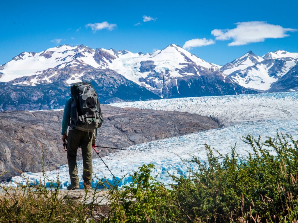 Un excursionista con una gran mochila y bastones de trekking contempla de espaldas la inmensidad del Glaciar Grey y las montañas nevadas de la cordillera de Paine en la Patagonia, una aventura desafiante para quienes deciden viajar solo en Chile.