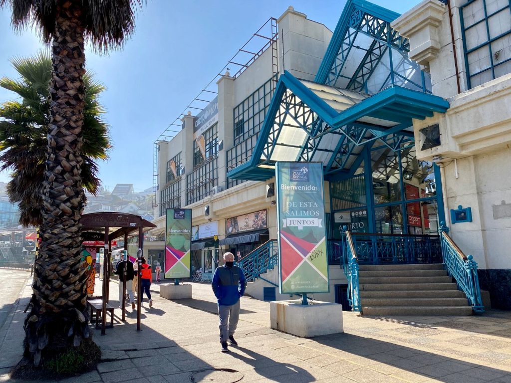 Fachada exterior del centro comercial Plaza Reñaca con sus estructuras y techos de color azul brillante, junto a una vereda con palmeras y peatones en un día soleado.