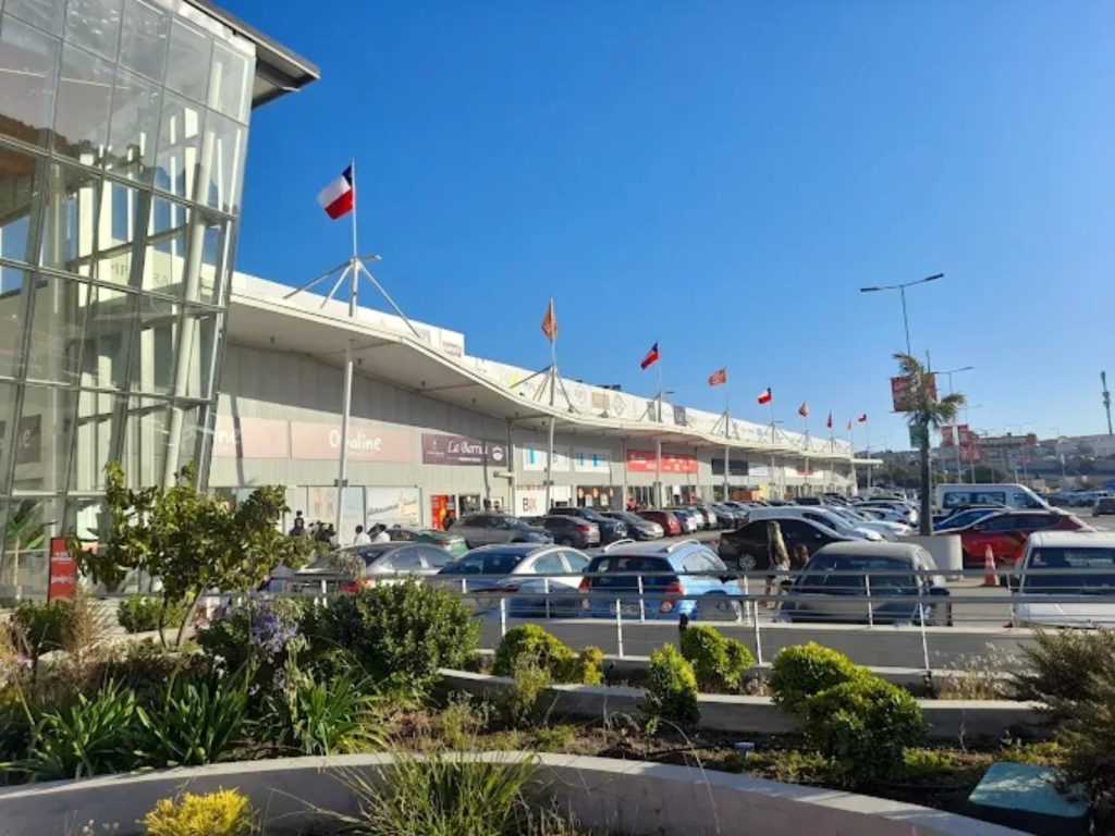 Estacionamiento exterior lleno de autos frente a la fachada de Viña Outlet Park decorado con banderas chilenas, bajo un cielo azul despejado.