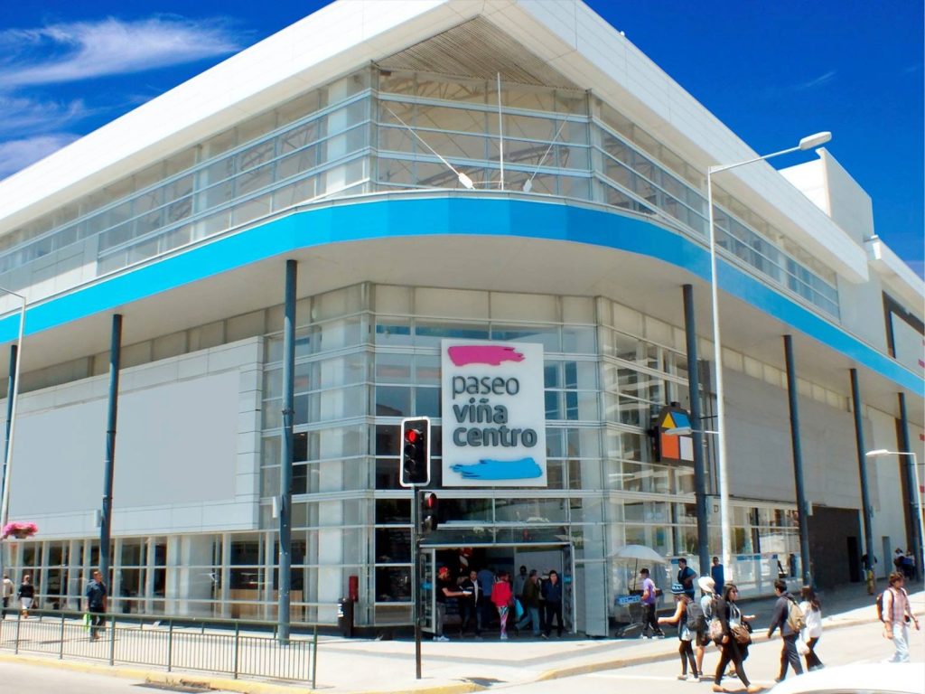 Fachada de esquina del centro comercial Paseo Viña Centro, destacando sus grandes ventanales de vidrio, pilares oscuros y un contorno azul brillante en la parte superior.
