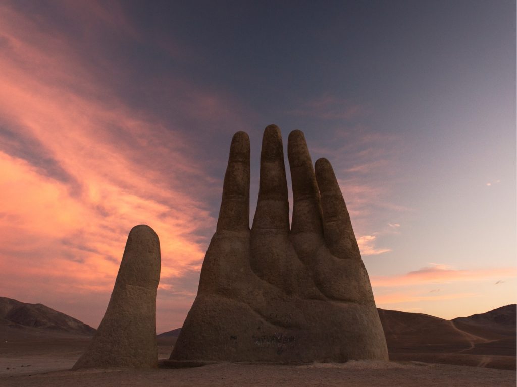 Silueta de la escultura de la Mano del Desierto bajo un cielo matutino con nubes rosadas al recorrer Chile por las extensas carreteras del norte.