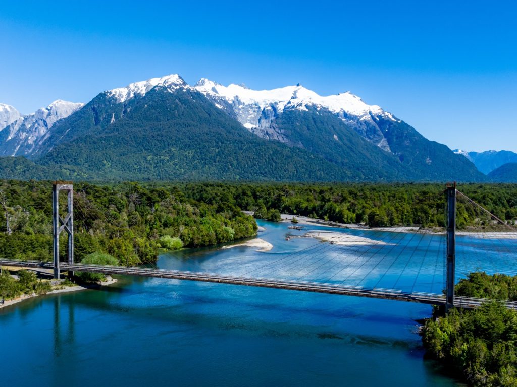 Vista aérea del puente colgante en la Carretera Austral rodeado de densos bosques y montañas nevadas, una ruta escénica clave al recorrer Chile.