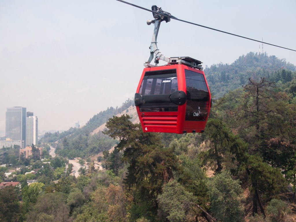 Una cabina roja del teleférico de Santiago avanza suspendida por cables en ell Cerro San Cristóbal, mostrando de fondo una vista parcial de la ciudad con edificios modernos bajo una bruma ligera, un panorama ideal al viajar solo en Chile.