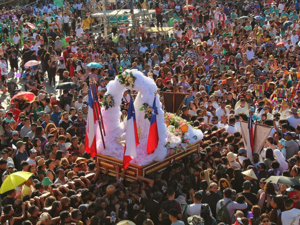 Vista aérea de una multitudinaria procesión de la Fiesta de La Tirana, donde una enorme masa de fieles rodea a la Virgen del Carmen, adornada con flores blancas y banderas chilenas en el Desierto de Atacama.