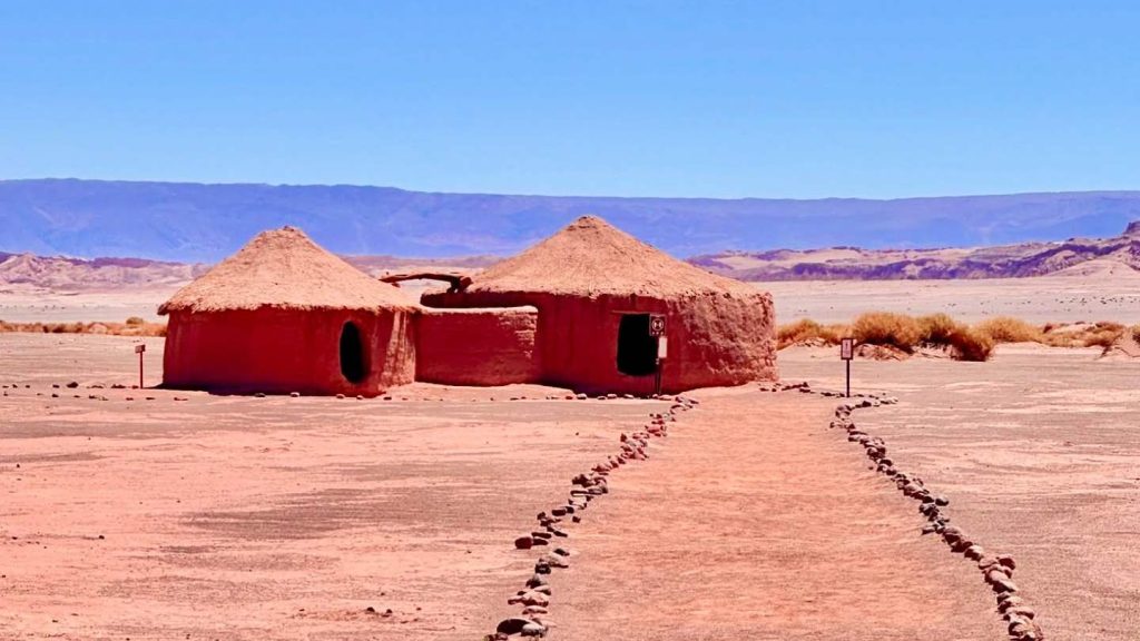 Close-up of two reconstructed circular dwellings with thick, rough-textured adobe walls and a conical roof made of mud and dry straw.