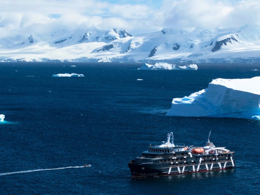 Un moderno crucero de expedición de color negro y blanco navega por las intensas aguas azules de la Antártica. Una de las aventuras más extremas al viajar a Chile. Al fondo se aprecian imponentes montañas cubiertas de nieve y grandes icebergs flotando cerca de la costa bajo un cielo parcialmente nublado.