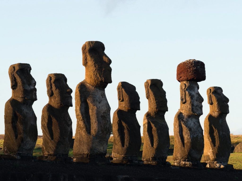 Una hilera de siete moái se alza sobre un ahu en Rapa Nui, uno de los destinos culturales más remotos de Chile. Las estatuas reciben la luz dorada del atardecer y una de ellas lleva su característico pukao de roca rojiza sobre la cabeza.