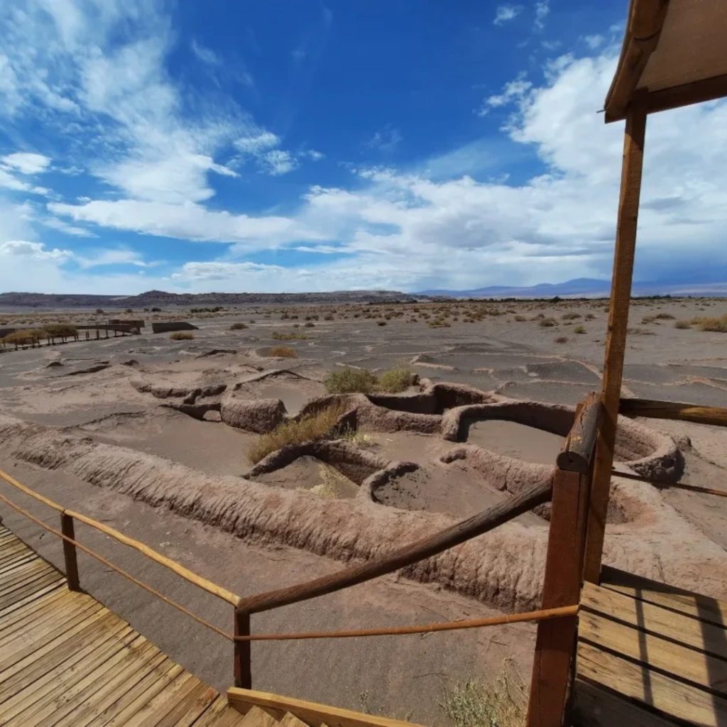From an elevated perspective on a wooden walkway and steps with log railings, the ancient circular mud foundations of Tulor Village are observed. The brown earth structures extend across the desert terrain, under a bright blue sky covered with dense clouds.