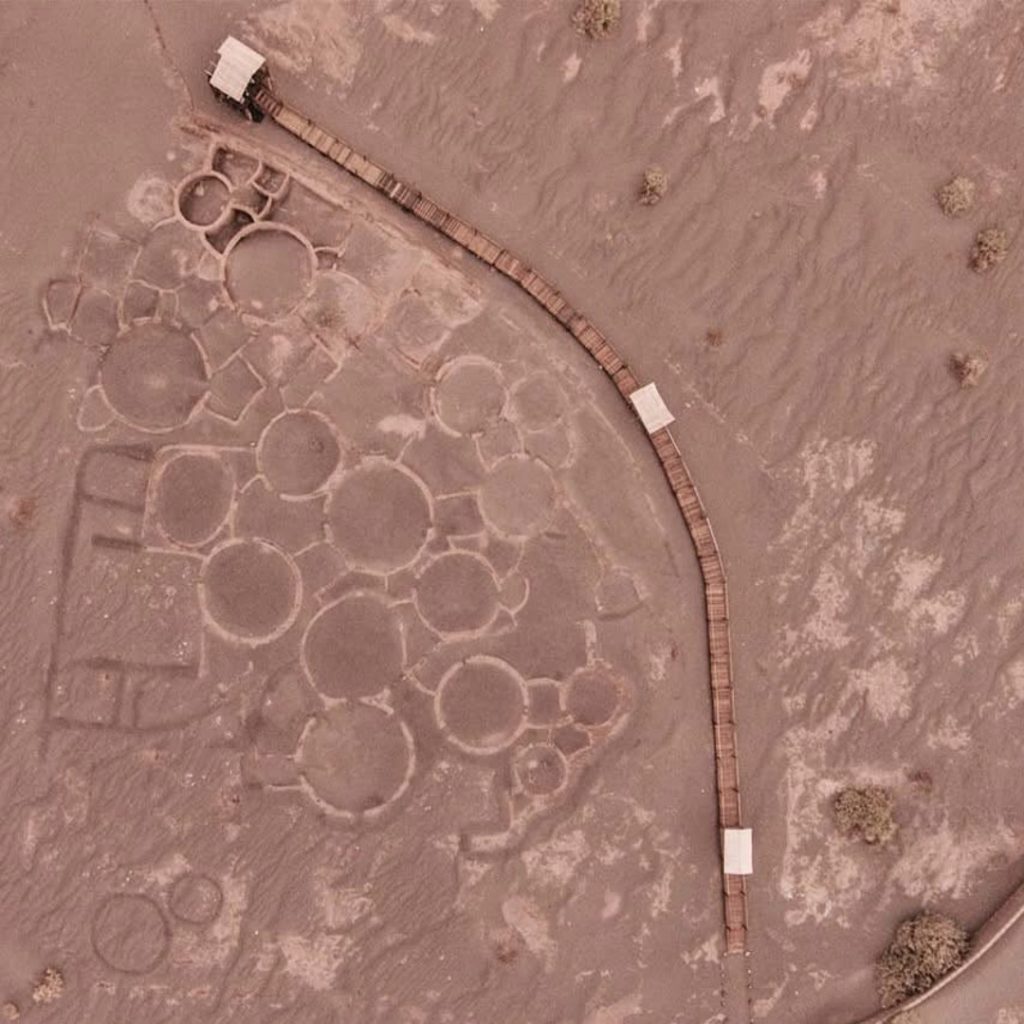 Aerial and overhead photograph showing the complete layout of Tulor Village. A cluster of multiple interconnected mud circles forming a labyrinthine pattern in the reddish-brown earth is visible.