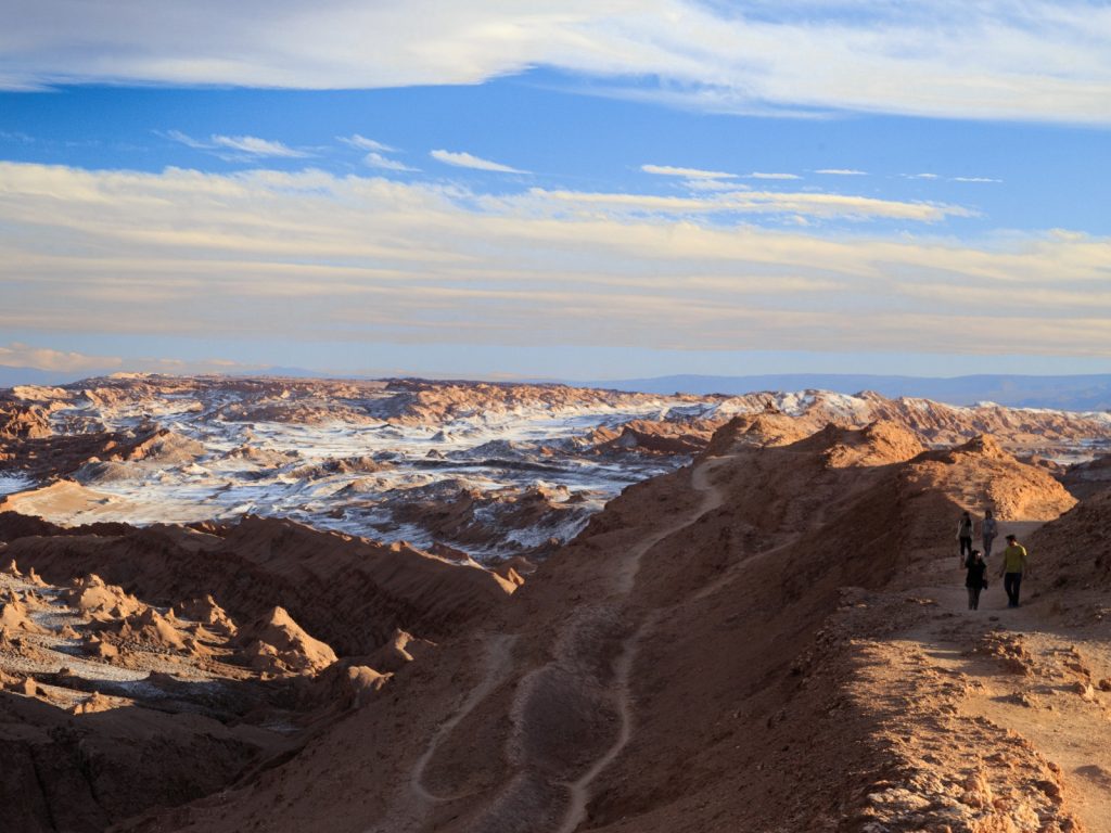 Un grupo de personas camina por un sendero en la cresta de una duna en el Valle de la Luna, mostrando los áridos e impresionantes paisajes del desierto de Atacama. El entorno destaca por sus formaciones rocosas rojizas texturizadas con rastros de sal blanca bajo un cielo azul con nubes ligeras.
