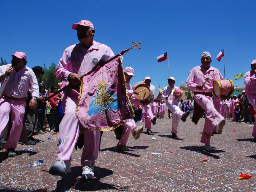 Un grupo de hombres pertenecientes a una cofradía de bailes chinos de Chile, vestidos completamente de rosa, tocan tambores y flautas mientras avanzan en procesión bajo un cielo azul despejado.