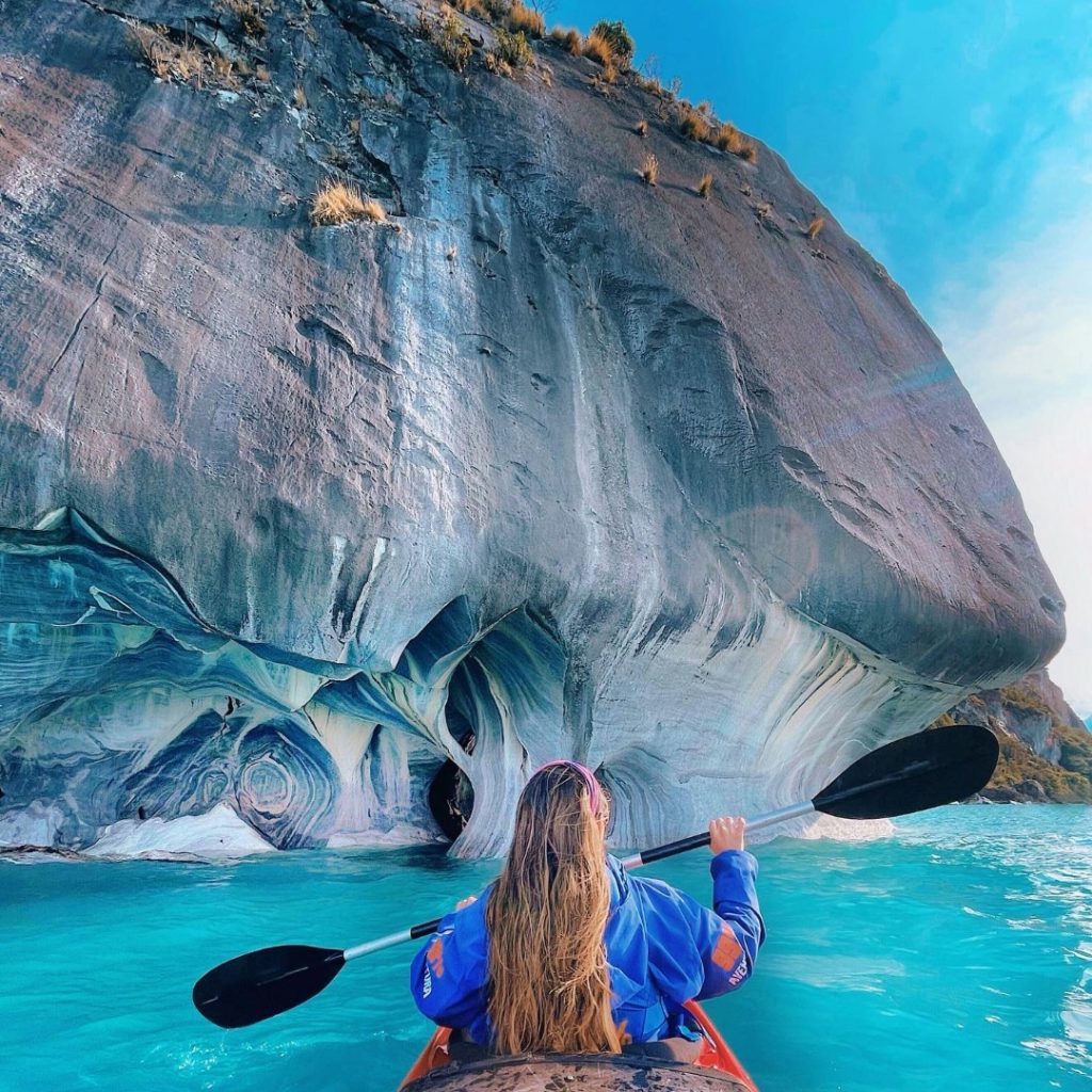 Una turista en kayak navegando frente a una enorme formación de las Capillas de Mármol, destacando los detallados patrones y vetas de la roca sobre el agua turquesa de la Patagonia.