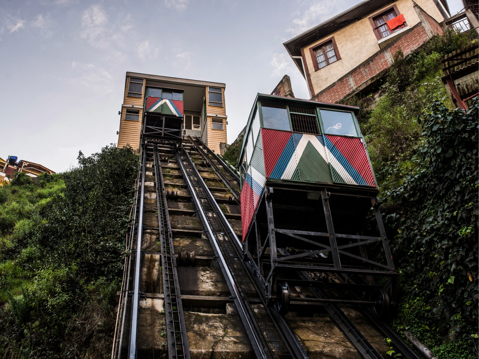 Ascensor histórico de Valparaíso com cabines coloridas a subir pelos seus carris entre os morros, rodeado de casas patrimoniais e vegetação nas encostas portenhas.