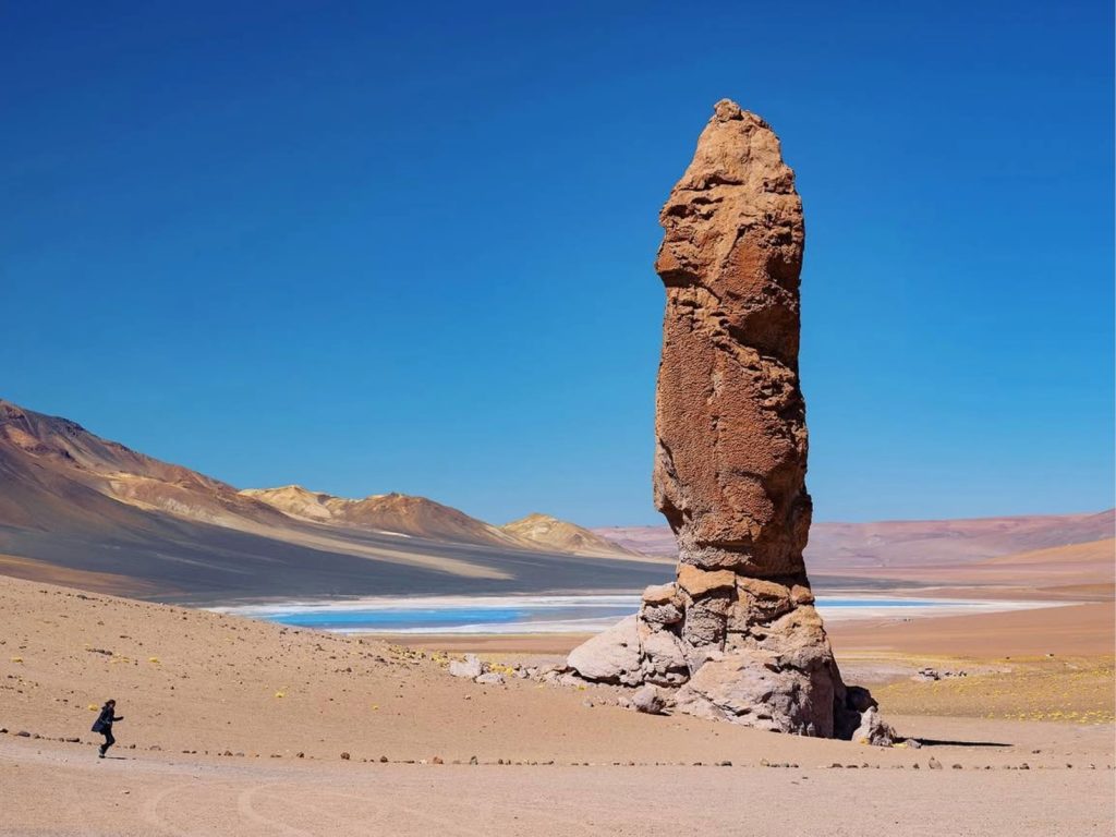 Imponente columna rocosa conocida como los Monjes de la Pacana, con una persona caminando a sus pies y una laguna turquesa junto a montañas coloridas de fondo, en el Salar de Tara, Región de Antofagasta.