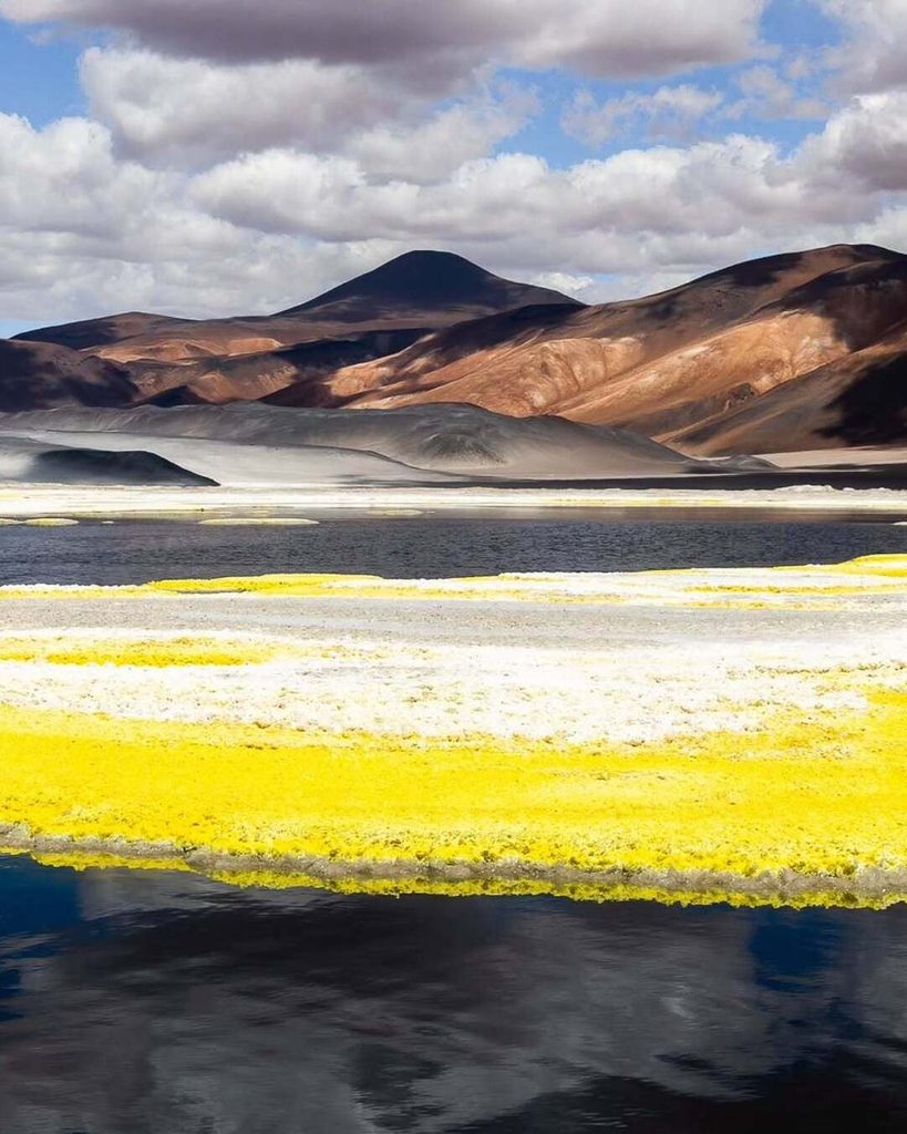 Laguna del Salar de Gorbea con una franja de sal en intensos tonos amarillos y blancos que se refleja en las aguas oscuras, rodeada de montañas multicolores, uno de los destinos secretos de Chile más sorprendentes del desierto de Atacama.