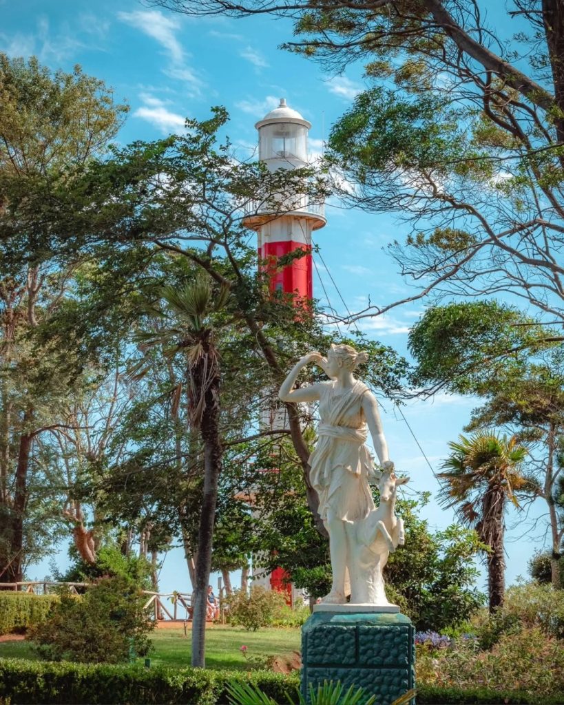 Estatua de mármol de una figura femenina junto a un ciervo, con un faro rojo y blanco asomando entre árboles y palmeras, en el Parque Isidora Cousiño de Lota.