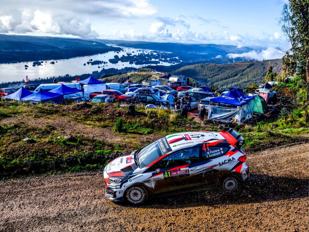 Auto de rally descendiendo por un camino de ripio en las alturas, con carpas de espectadores y un lago rodeado de bosques al fondo, uno de los tramos de montaña del rally.