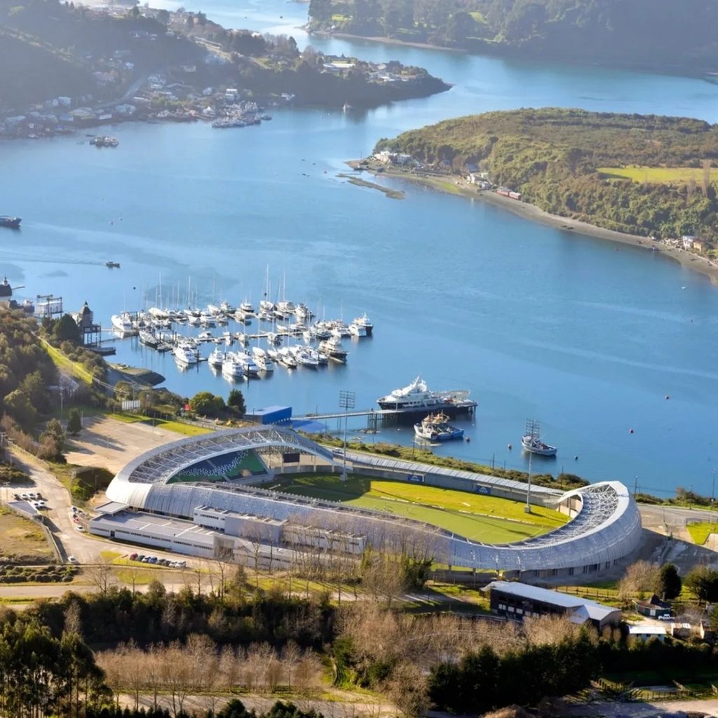 Vista aérea del Estadio Bicentenario Chinquihue en Puerto Montt, frente al Seno de Reloncaví con embarcaciones ancladas y cerros boscosos al fondo.