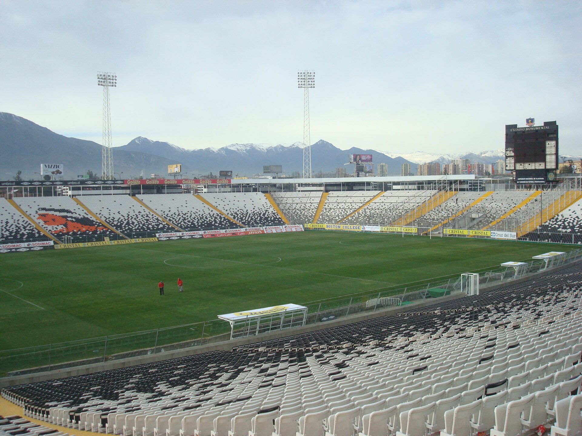 Tribünen des Estadio Monumental mit dem Mosaik des Colo-Colo-Emblems, das in den Sitzen geformt ist, Sponsorenwerbung und die schneebedeckten Anden im Hintergrund.
