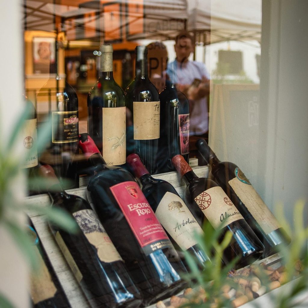 Vitrina con botellas de vinos chilenos antiguos de distintas viñas en Barrio Lastarria, recostadas sobre repisas de madera y rodeadas de corchos, con el reflejo de la calle en el vidrio.