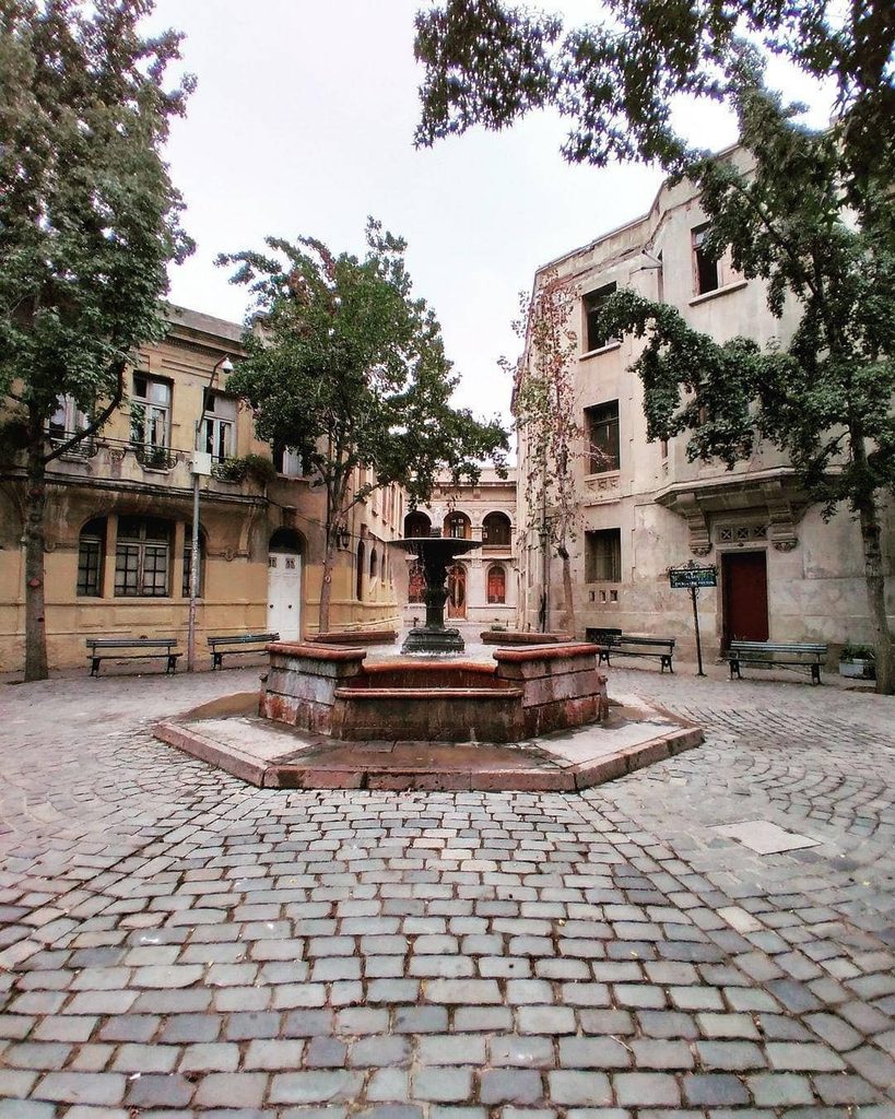 Pileta de piedra en la plaza adoquinada del Barrio Concha y Toro en Santiago, rodeada de árboles, bancas y fachadas de casonas de principios del siglo XX