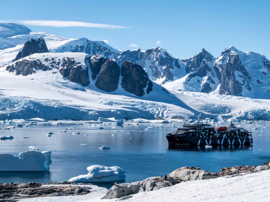 Barco de expedición navegando entre glaciares e icebergs, rodeado de montañas nevadas en la Antártica.