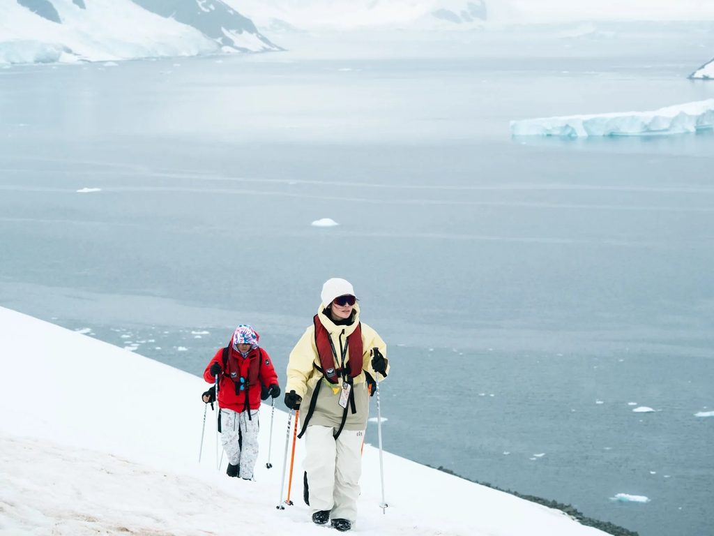 Pasajeros con bastones de trekking caminando sobre la nieve frente al mar, durante una excursión en la Península Antártica.