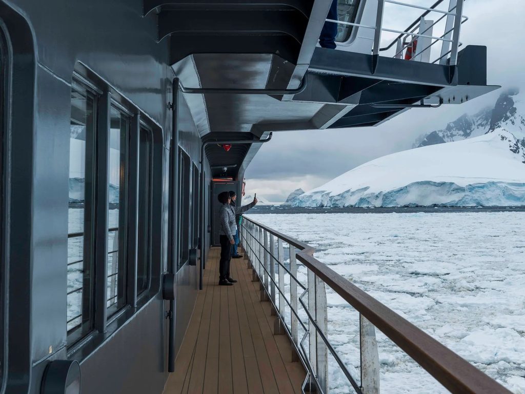 Pasajeros de un crucero a la Antártica observando icebergs y montañas nevadas desde la cubierta de la embarcación.