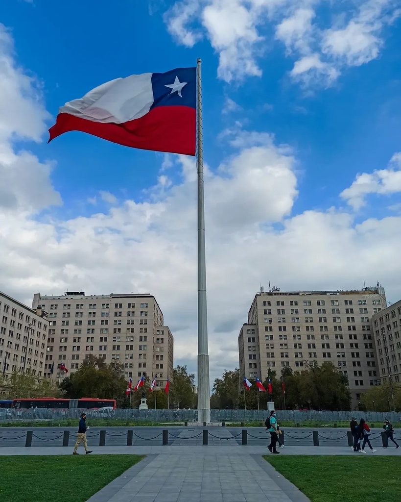 Gran Bandera Bicentenario ondeando sobre un mástil en el Barrio Cívico de Santiago, con edificios de fondo.