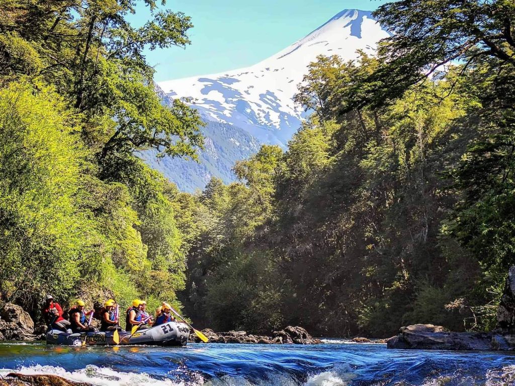 Grupo haciendo rafting en un río rodeado de bosque nativo, con un volcán nevado al fondo en los alrededores de Pucón.