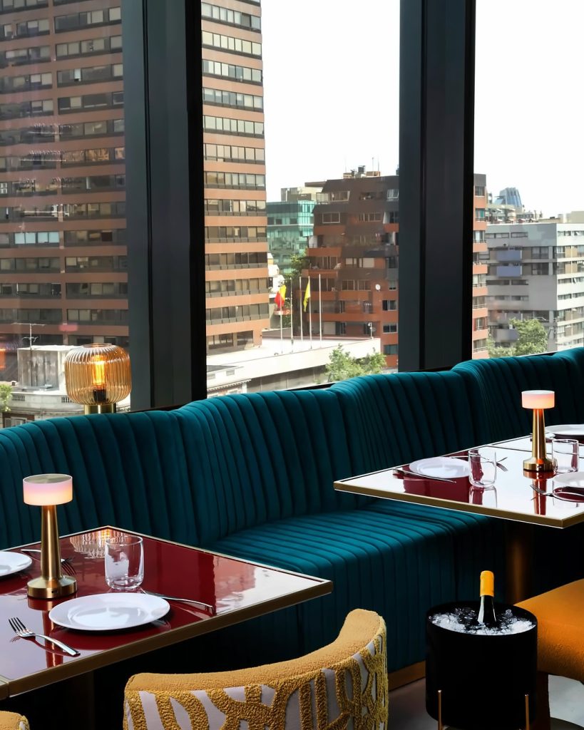 Sillones de terciopelo verde, mesas lacadas y lámparas doradas frente a los ventanales con vista a Santiago en The Loft, un bistró mediterráneo.