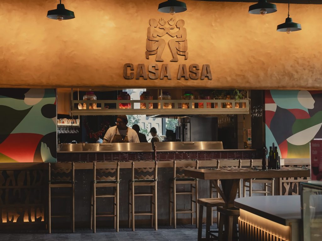 Barra de madera y cocina abierta bajo el logo de Casa Asa, restaurante de preparaciones a las brasas para compartir.