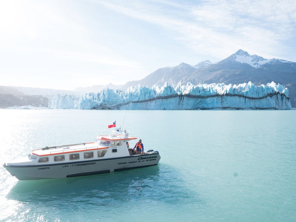 Lancha de turismo navegando por las aguas turquesa frente al glaciar O’Higgins y montañas nevadas, en Villa O’Higgins, Chile.