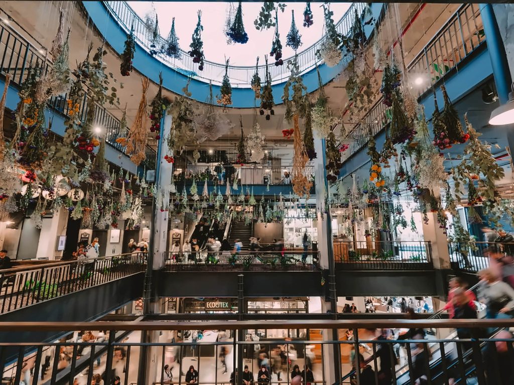 Instalación de flores colgantes bajo la claraboya del MUT, en Santiago, con varios niveles de tiendas y visitantes recorriendo el mercado.