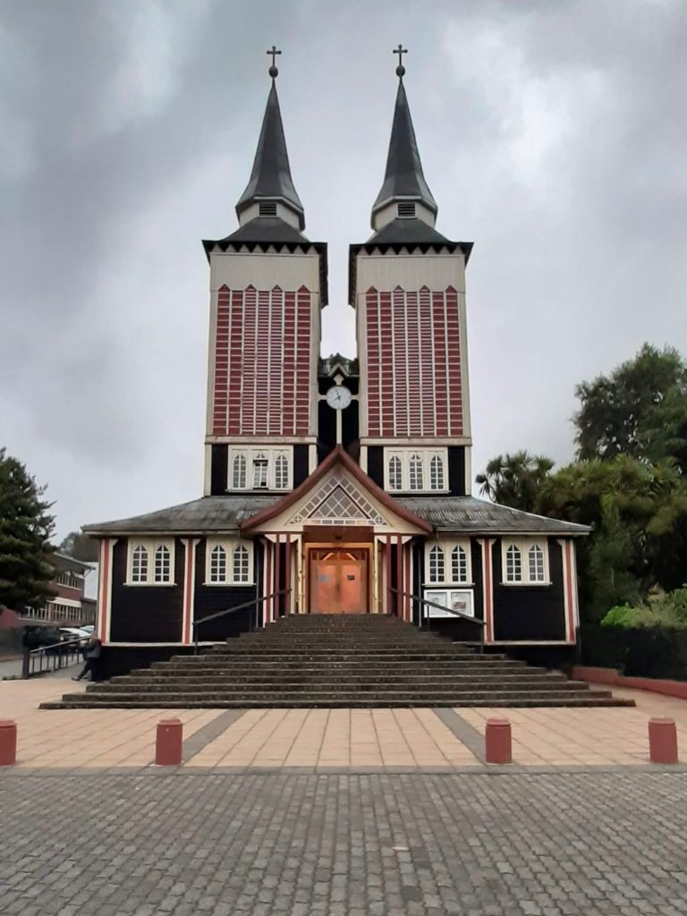 Iglesia San Sebastián de Panguipulli vista de frente con su distintiva estructura de madera, escalinata de acceso y dos imponentes torres simétricas rematadas en agujas con cruces. Destacan sus detalles en tonos rojo, blanco y café bajo un cielo nublado característico del sur de Chile.
