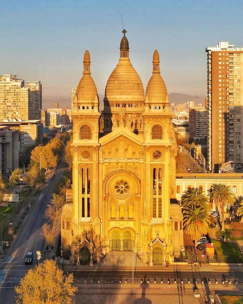 Basílica del Santísimo Sacramento iluminada por la luz dorada del atardecer. Templo de gran escala con cúpulas de estilo bizantino-románico, torres simétricas y rosetón central. A los lados se observan avenidas, edificios urbanos y palmeras bajo la cálida luz solar.