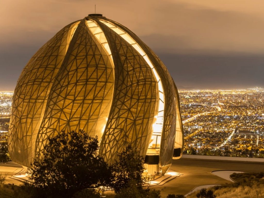 Templo Bahá'í de Sudamérica en Peñalolén iluminado durante la noche con su moderna estructura en forma de flor o capullo. Al fondo se aprecia una panorámica vista nocturna de la ciudad de Santiago.