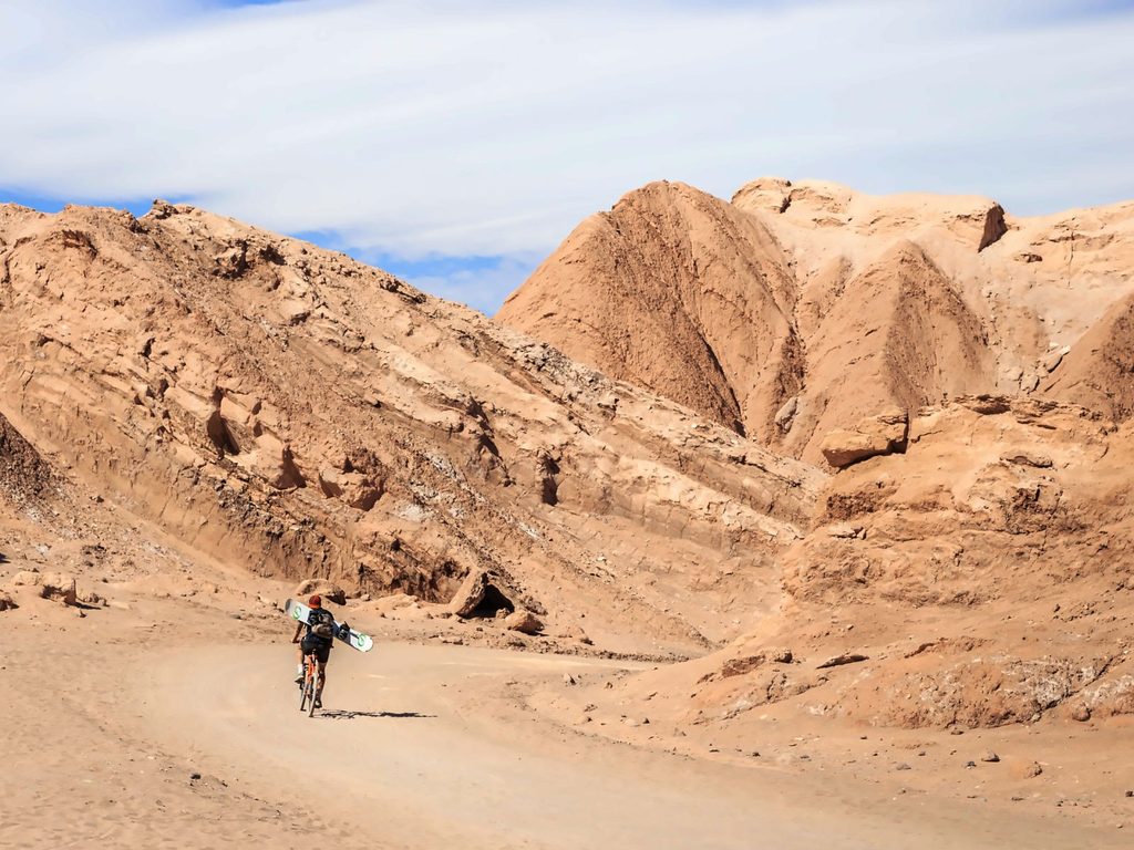 Ciclista con una tabla de sandboard recorriendo un camino entre cerros de arcilla en el Valle de la Muerte, en San Pedro de Atacama.