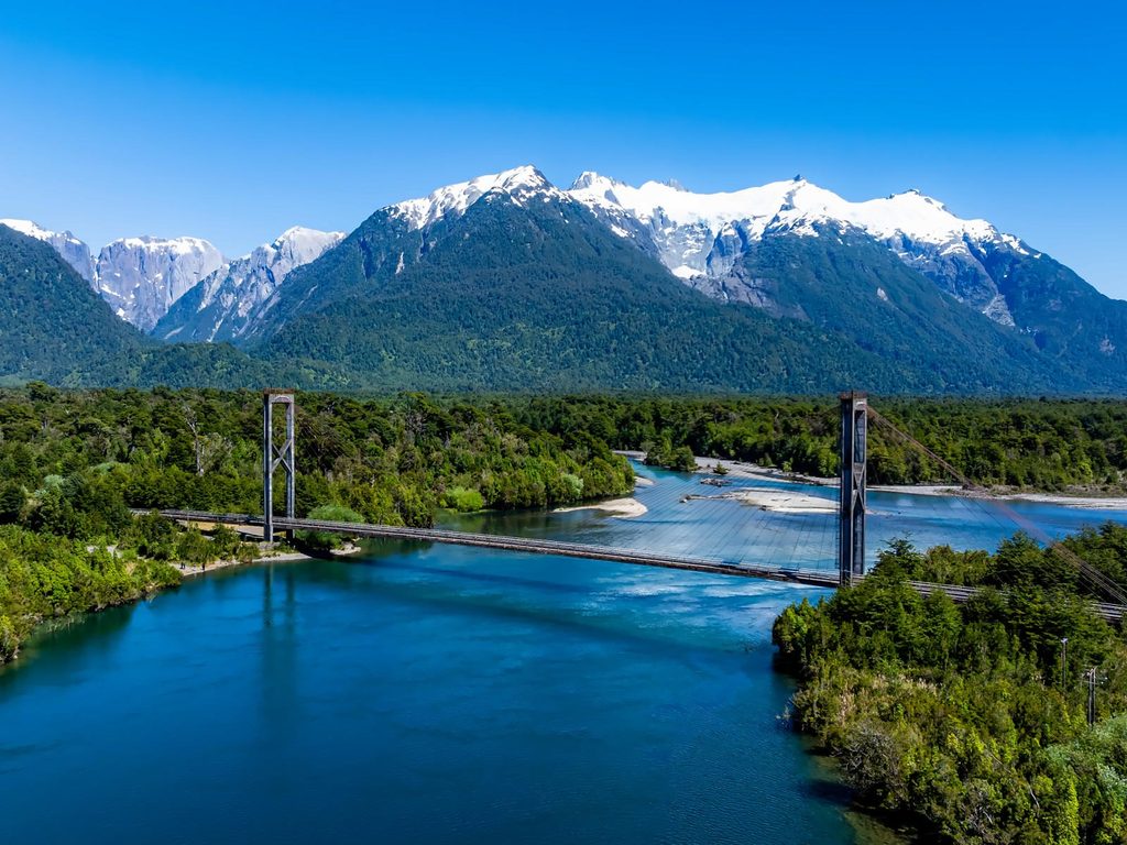 Puente colgante sobre un río de aguas azules rodeado de bosque y montañas nevadas en la Carretera Austral.