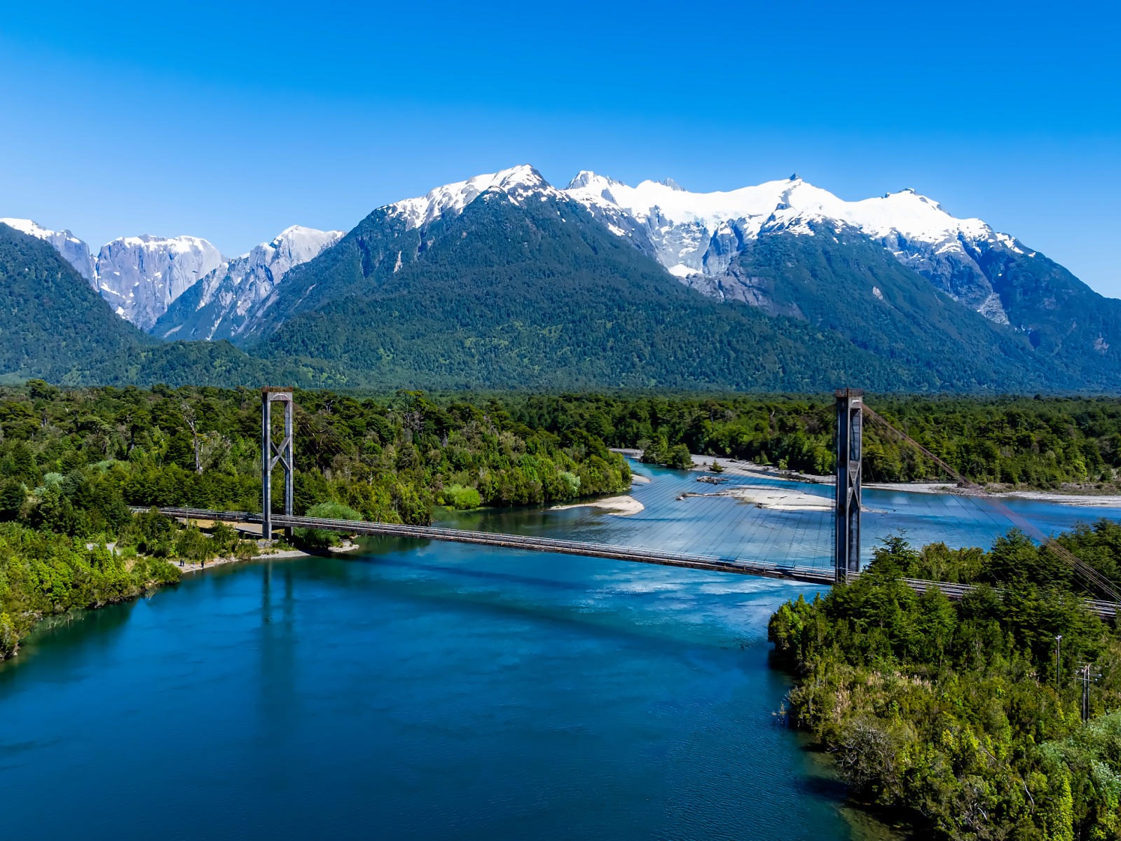 Suspension bridge over a blue-water river surrounded by forest and snow-capped mountains on the Carretera Austral.