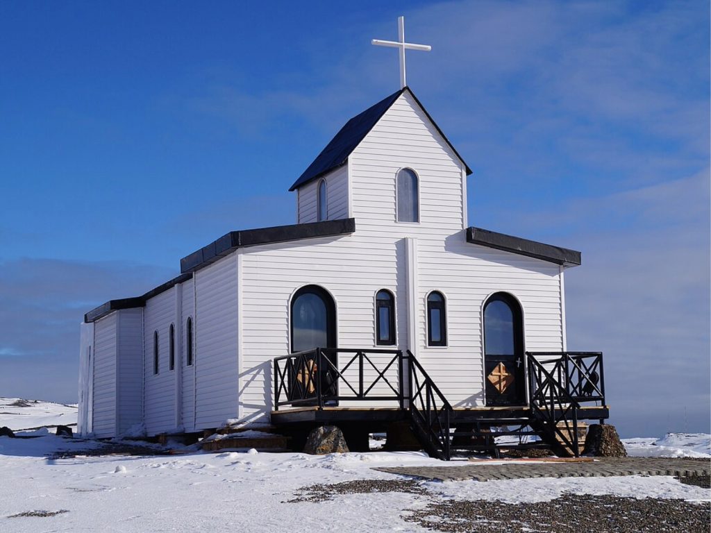 Capilla de Santa María Reina de la Paz en la Antártica Chilena vista de frente con su fachada de madera blanca y detalles oscuros en las barandillas y tejados, coronada por una cruz blanca en el pico del techo.