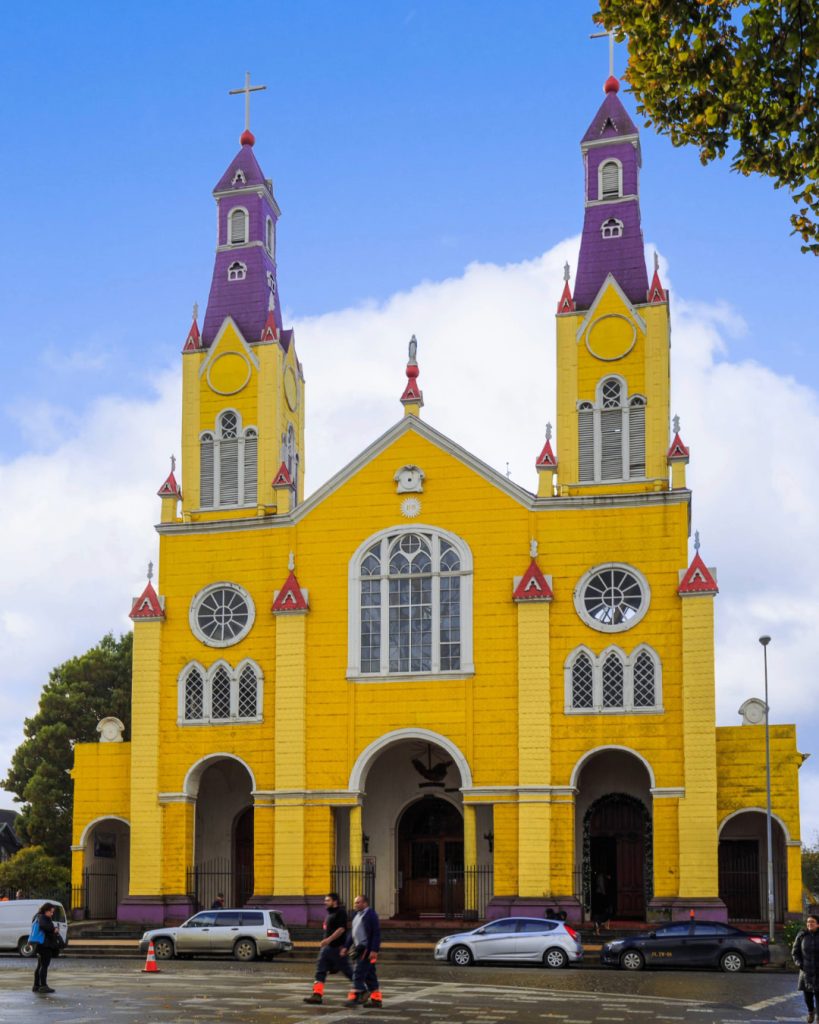 Iglesia San Francisco de Castro vista de frente con sus fachadas en amarillo y detalles en morado. Destacan sus dos torres simétricas rematadas en agujas con cruces, sus arcos de acceso y ventanales con rosetones bajo un cielo parcialmente nublado.