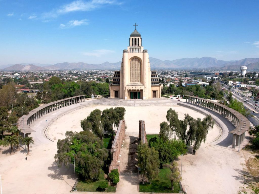 Vista aérea del Templo Votivo de Maipú con su imponente estructura monumental coronada por una cruz. En el primer plano destaca la amplia explanada semicircular, rodeada de columnatas, áreas verdes y los muros históricos en ruinas bajo un cielo azul despejado.